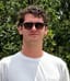 Blake Deutser wearing sunglasses and a white T-shirt, standing outdoors in front of green foliage.